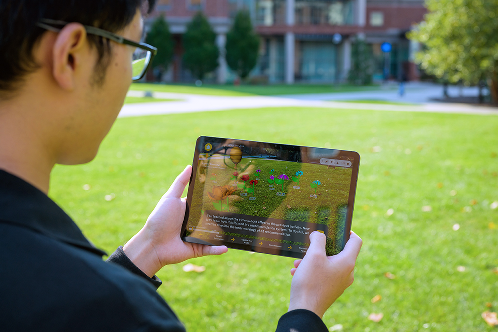 A person holding a tablet in front of them on the Hajim Quad with a building in the background.