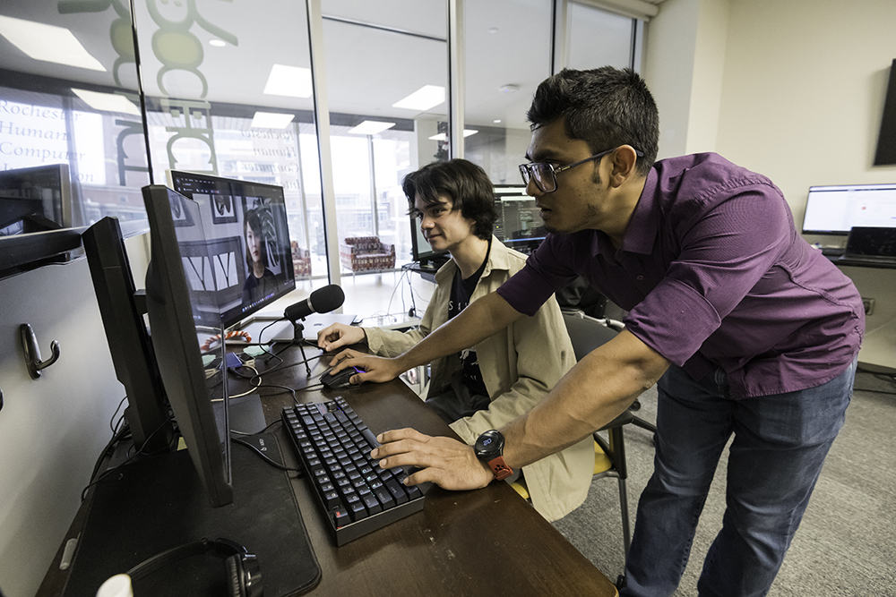 A computer science student (seated) interacts with SAPIEN, an avatar built by the team as a PhD student looks on.