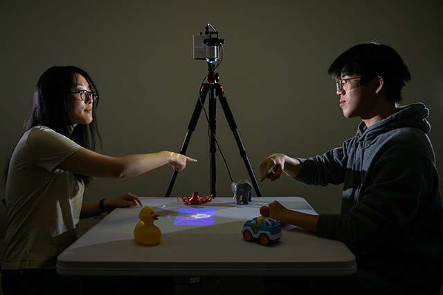 Two students face each other across a table in a darkened room with some objects and a video recorder.